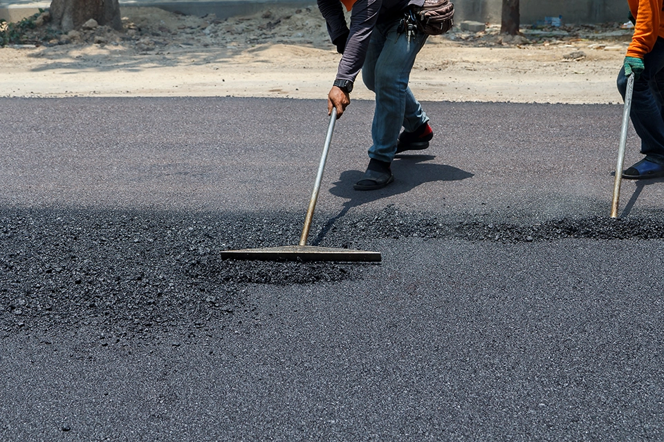 A crew member operates a paving machine, smoothing hot asphalt on a construction site.